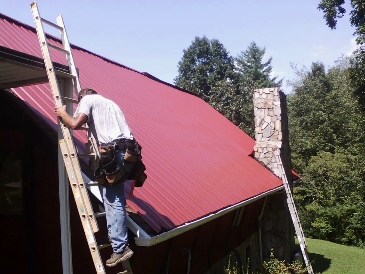 Roofer on a ladder beside a red metal roof and stone chimney — Soffit & Fascia Repair by Roofing Brothers in Shelburne Falls