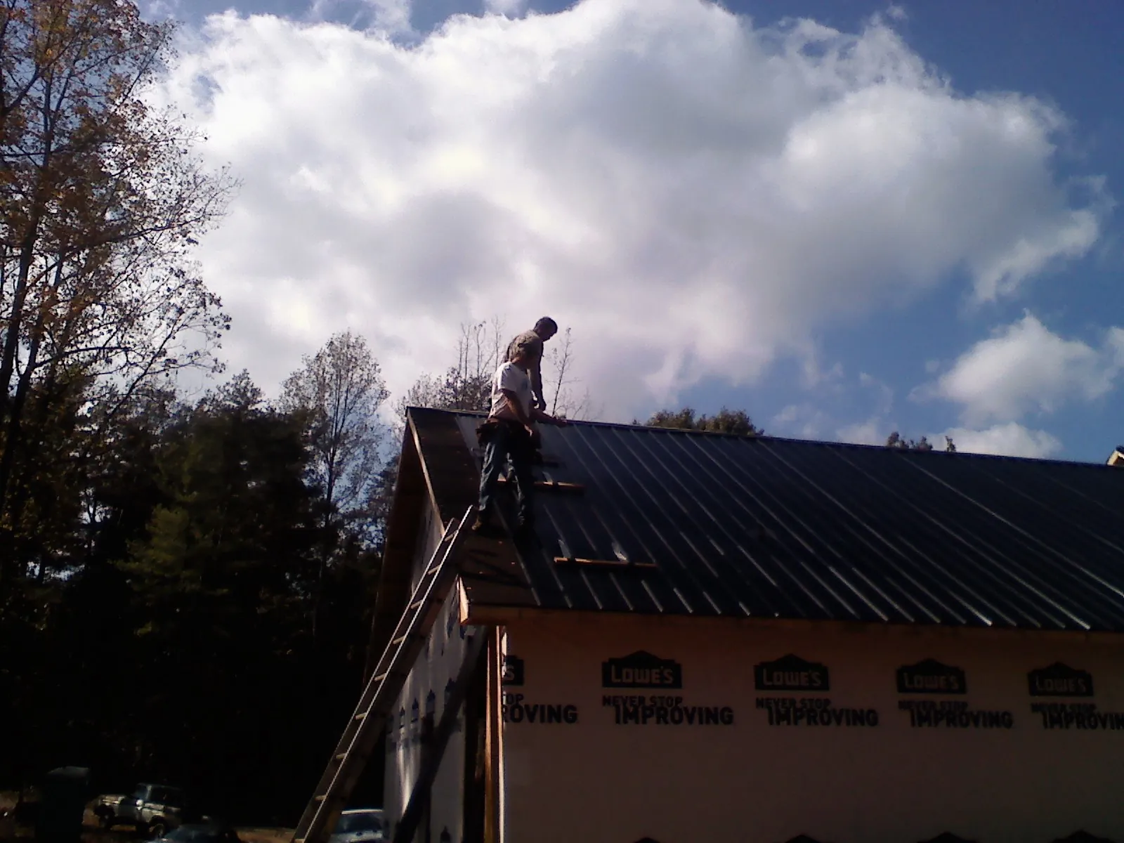 Roofer fastening dark metal panels high against a cloudy sky, from a Roof Inspection job in Shelburne Falls